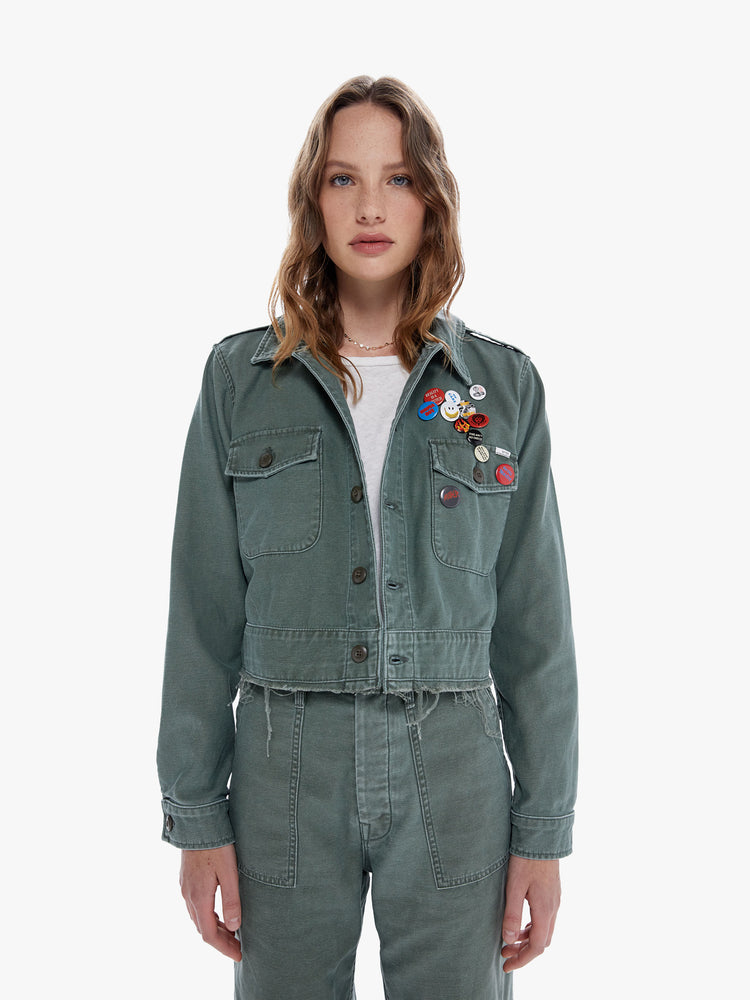 Front close up view of a woman in a military inspired jacket with a front patch pockets and a cropped, fray hem made from 100% cotton deadstock fabric, Roger That is a classic army green hue with tonal buttons and decorative pins on the chest
