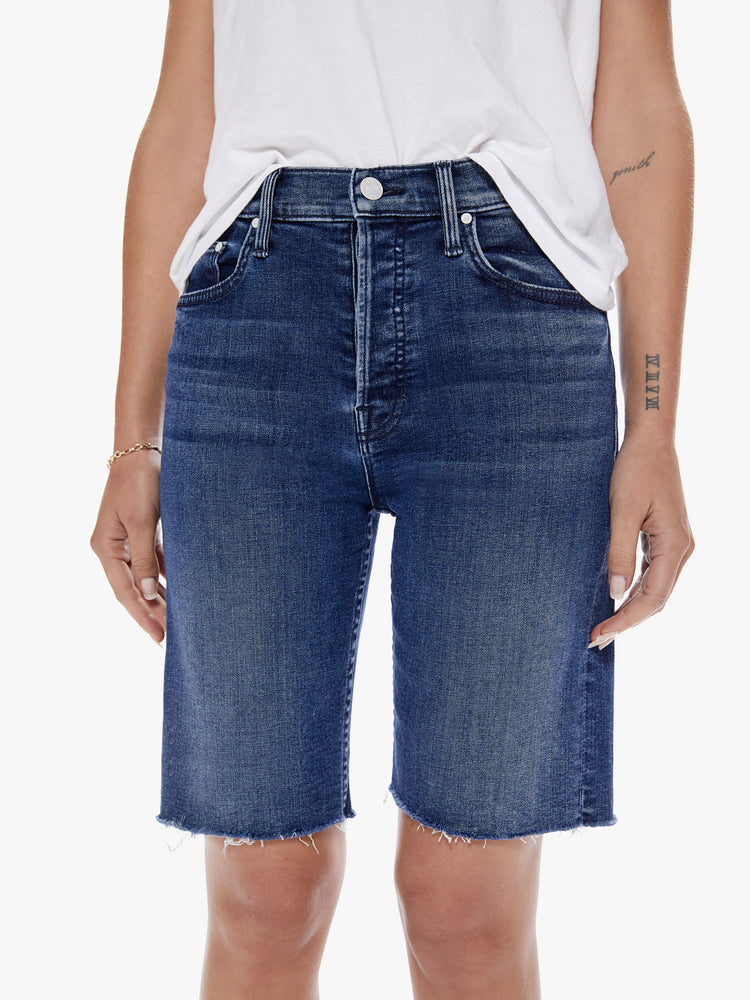 Waist full body view of a woman in a knee-length denim shorts with a high rise, button fly and frayed hem cut from a blend of organic cotton with a touch stretch in an indigo-blue wash with subtle whiskering and fading