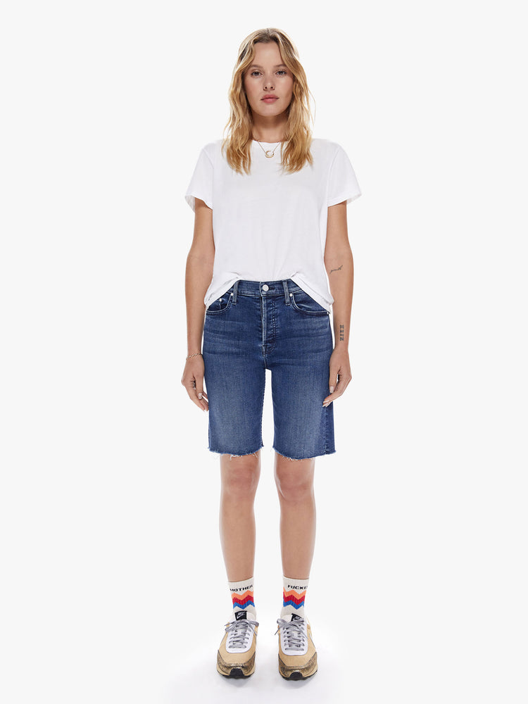 Front full body view of a woman in a knee-length denim shorts with a high rise, button fly and frayed hem cut from a blend of organic cotton with a touch stretch in an indigo-blue wash with subtle whiskering and fading