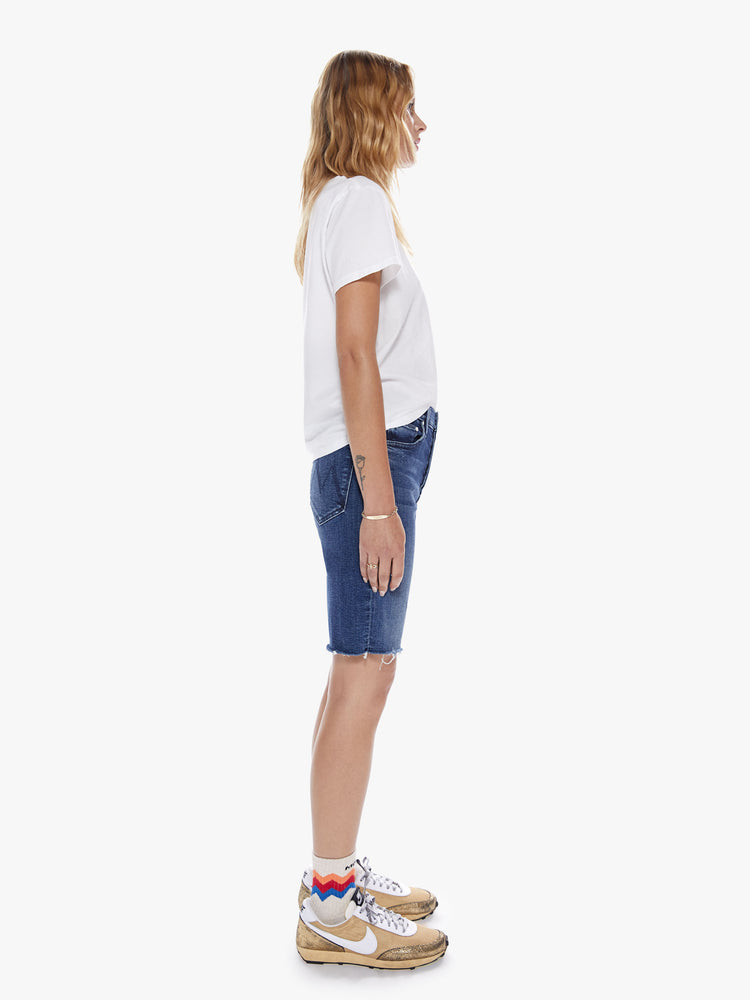 Side full body view of a woman in a knee-length denim shorts with a high rise, button fly and frayed hem cut from a blend of organic cotton with a touch stretch in an indigo-blue wash with subtle whiskering and fading
