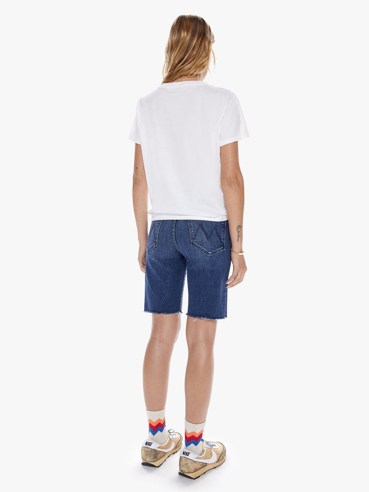 Back full body view of a woman in a knee-length denim shorts with a high rise, button fly and frayed hem cut from a blend of organic cotton with a touch stretch in an indigo-blue wash with subtle whiskering and fading