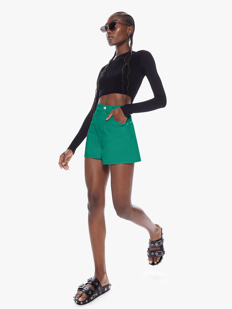 Front in motion full body view of a woman in denim shorts from SNACKS from mothers homage to throwback styles of the 80s and 90s, the high rise shorts are designed with a super short inseam and a loose wide fit in a forest green hue