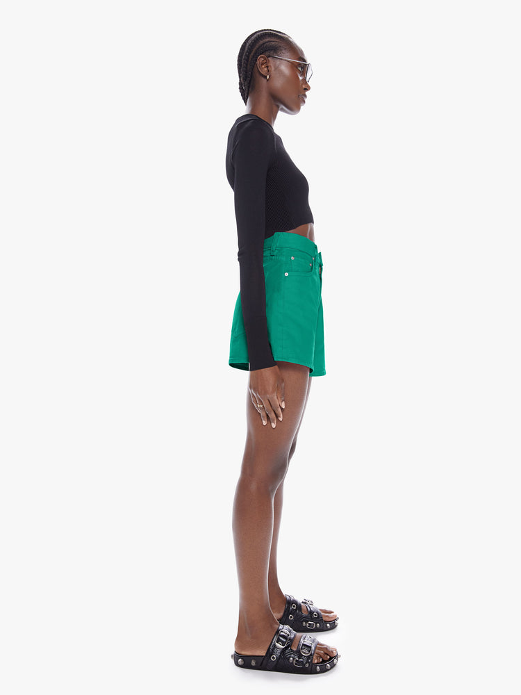 Side full body view of a woman in denim shorts from SNACKS from mothers homage to throwback styles of the 80s and 90s, the high rise shorts are designed with a super short inseam and a loose wide fit in a forest green hue