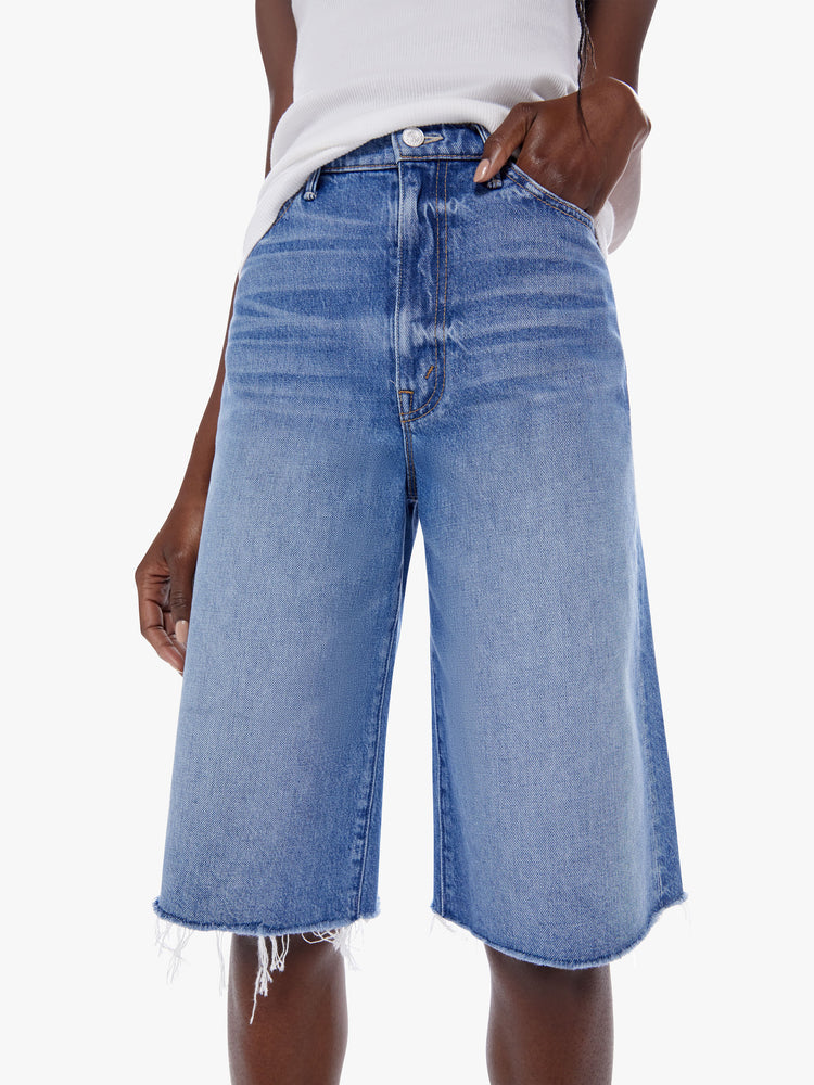 Close up view of a woman knee-length shorts feature a high rise, wide leg and frayed hem in a mid blue wash.