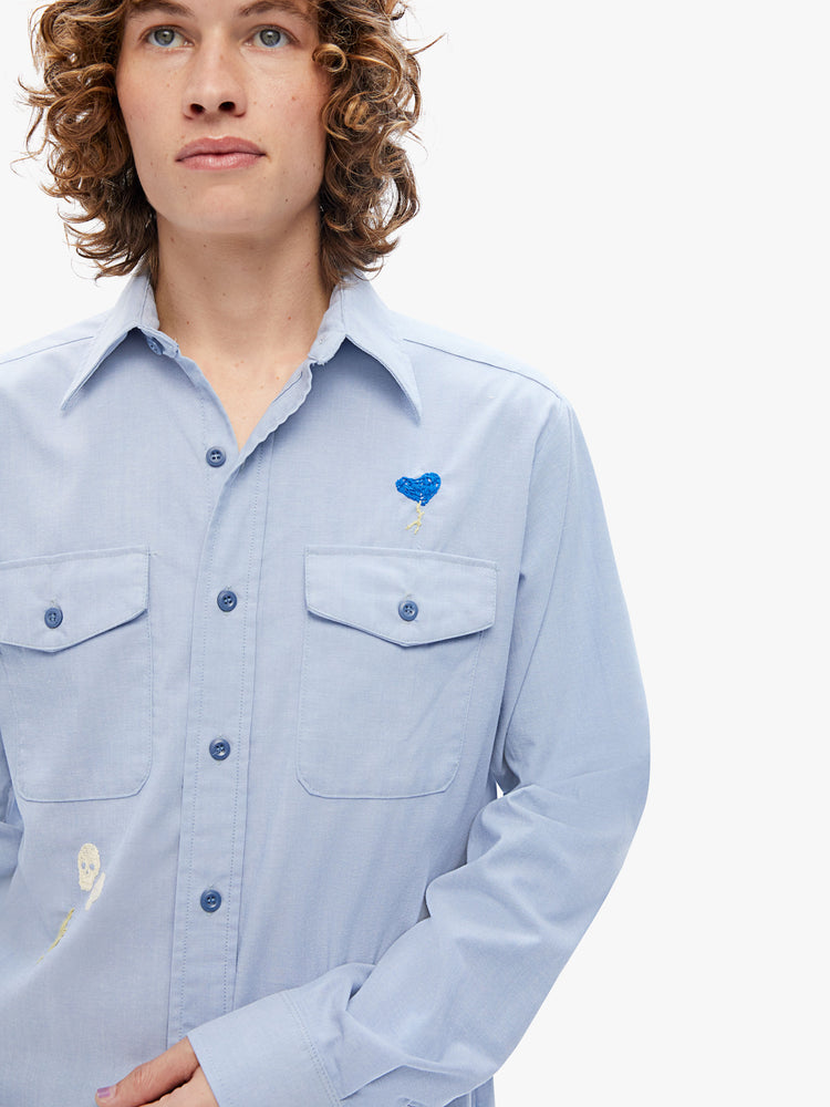 Close up view of a mens long-sleeve button-up with front patch pockets and a curved hem in a light blue hue with embroidered details on the front and back.