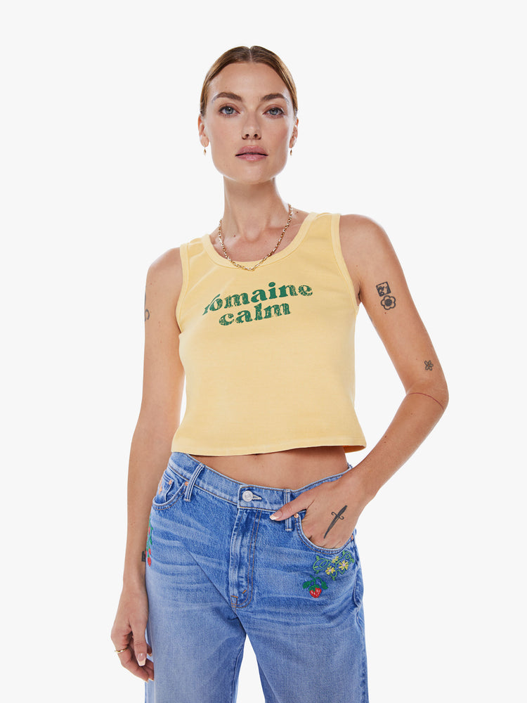 Front view of woman tank with a crewneck, thick straps and a cropped hem in a pastel yellow hue with green text.