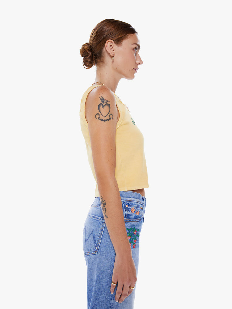Side view of woman tank with a crewneck, thick straps and a cropped hem in a pastel yellow hue with green text.