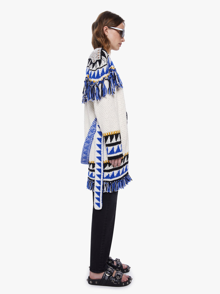 Side view of a womens black and blue triangle print thigh length cardigan sweater with orange knit details, tassels across the chest, arms, hems and a slight dropped shoulders and a boxy oversized fit