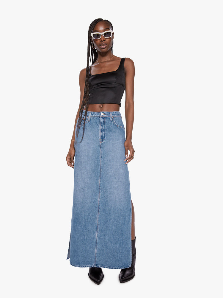 Front view of a woman wearing a medium blue wash denim maxi skirt featuring a mid rise and high side slits.