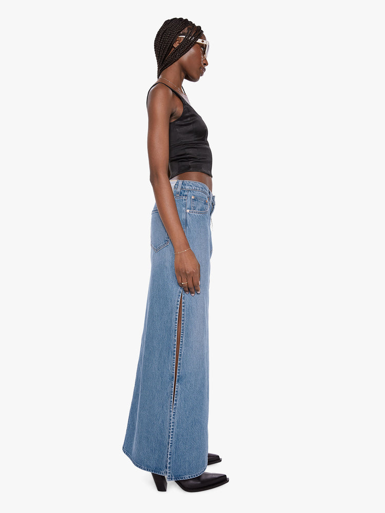 Side view of a woman wearing a medium blue wash denim maxi skirt featuring a mid rise and high side slits.