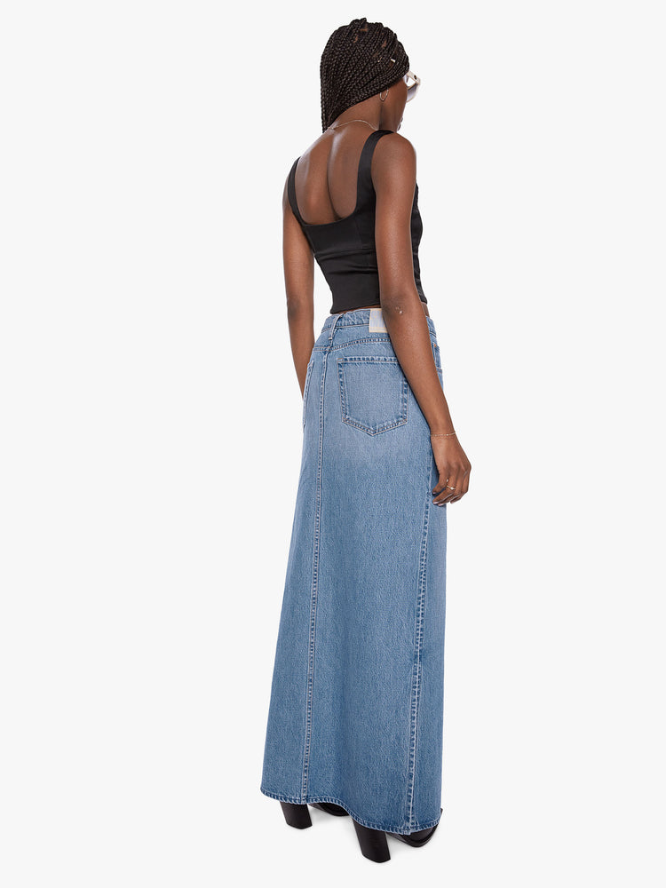 Back view of a woman wearing a medium blue wash denim maxi skirt featuring a mid rise and high side slits.