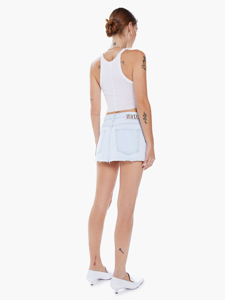 Back view of woman denim mini skirt worn loose and low at the hips, the skirt features a zip fly, exposed pocket liners and a raw hem in a faded light blue wash.