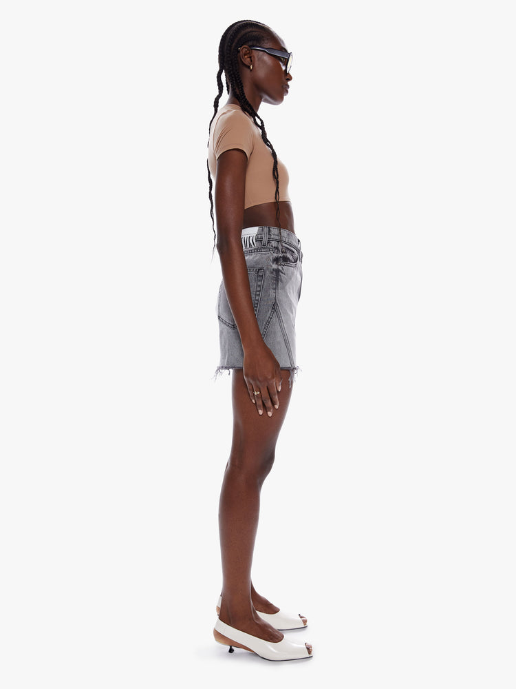 Side full body view of a woman in a denim mini skirt from snacks Mother homage to throwback styles of the 80s and 90s, this high rise denim skirt features a short frayed hem and a snug fit in a washed grey hue