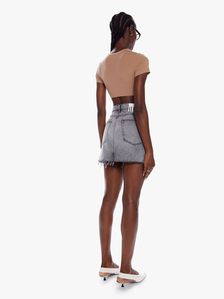 Back full body view of a woman in a denim mini skirt from snacks Mother homage to throwback styles of the 80s and 90s, this high rise denim skirt features a short frayed hem and a snug fit in a washed grey hue