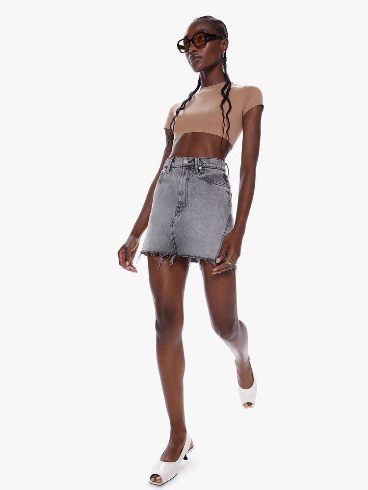 In motion full body view of a woman in a denim mini skirt from snacks Mother homage to throwback styles of the 80s and 90s, this high rise denim skirt features a short frayed hem and a snug fit in a washed grey hue