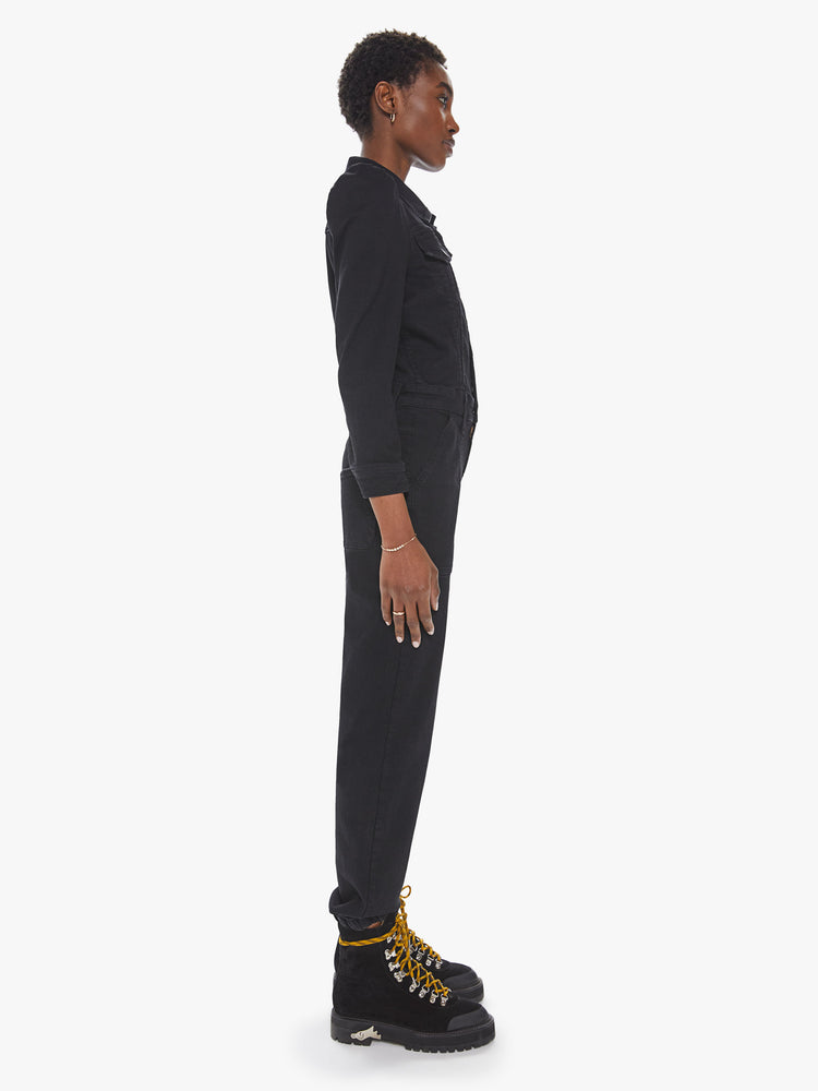 Side view of a womens black wash denim jumpsuit featuring a button down collar, front chest pockets, cinched waist, long sleeves, and an elastic pant hem..