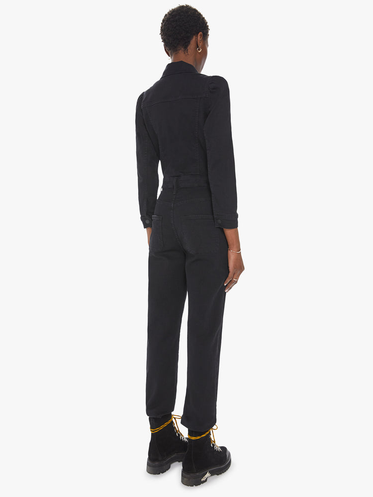 Back view of a womens black wash denim jumpsuit featuring a button down collar, front chest pockets, cinched waist, long sleeves, and an elastic pant hem..