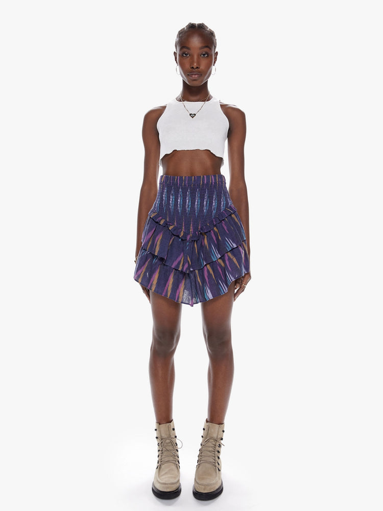 Front full body view of a woman in a high rise skirt with a smocked elastic waistband, v-shapped ruffles and playfully short hem made from 100% cotton in a dark blue hue with a brush stroke-inspired pattern in lavender, mustard, and baby blue