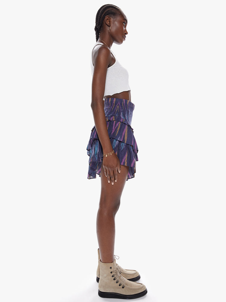 Side  full body view of a woman in a high rise skirt with a smocked elastic waistband, v-shapped ruffles and playfully short hem made from 100% cotton in a dark blue hue with a brush stroke-inspired pattern in lavender, mustard, and baby blue