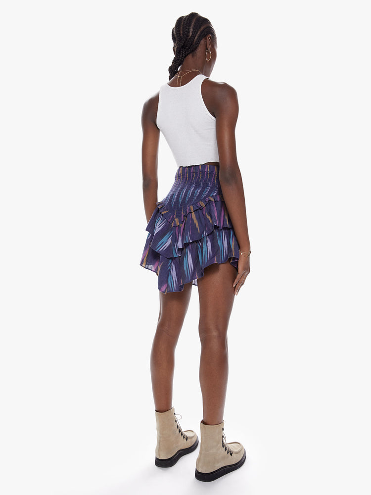 Back  full body view of a woman in a high rise skirt with a smocked elastic waistband, v-shapped ruffles and playfully short hem made from 100% cotton in a dark blue hue with a brush stroke-inspired pattern in lavender, mustard, and baby blue