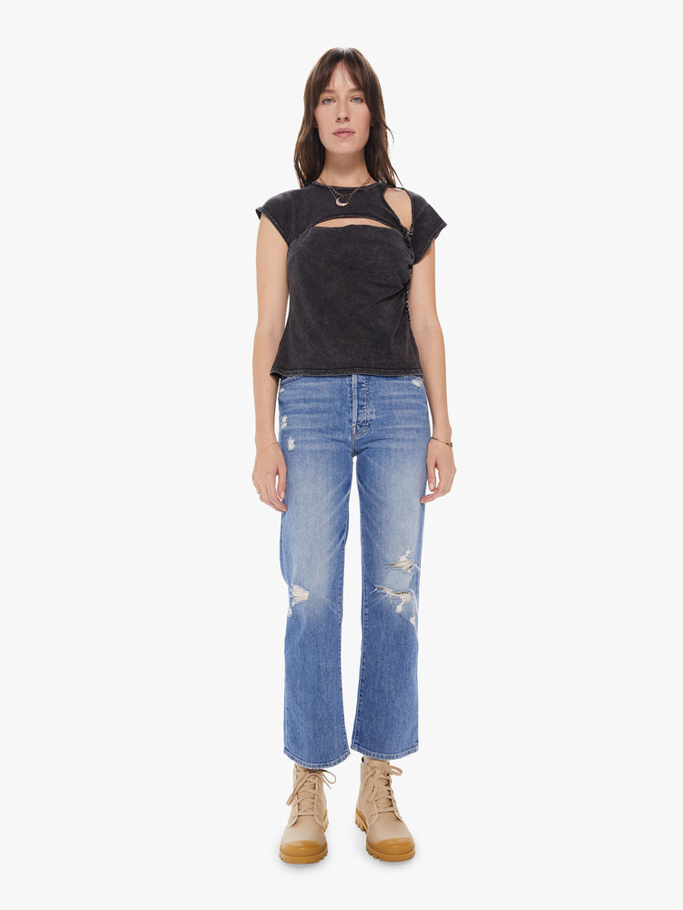 A full body view of a Womens asymmetrical sleeve top in a black mineral wash, featuring cutouts at the shoulder and chest and a braid down the left side, paired with a mid blue wash straight leg jean.