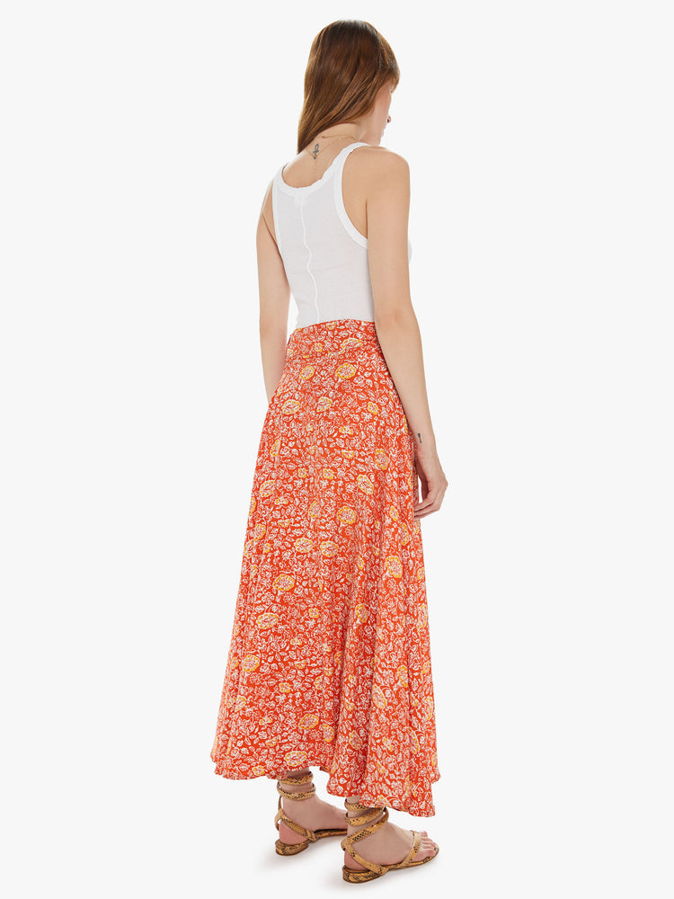 Back view of a woman wearing a bright orange maxi skirt featuring a yellow print, a high waist, and a side wrap tie.