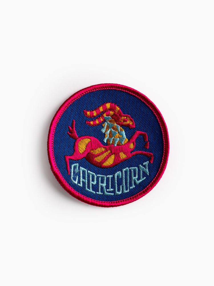 A top front view of a round, blue embroidered patch with contrast pink stitching, featuring a colorful sea goat with the words "CAPRICORN"