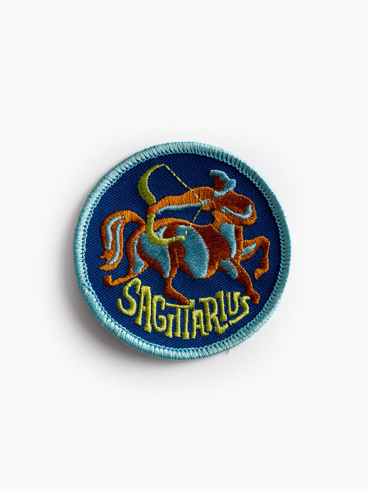 A top front view of a round, blue embroidered patch with contrast light blue stitching, featuring a colorful archer and the word "SAGITTARIUS"