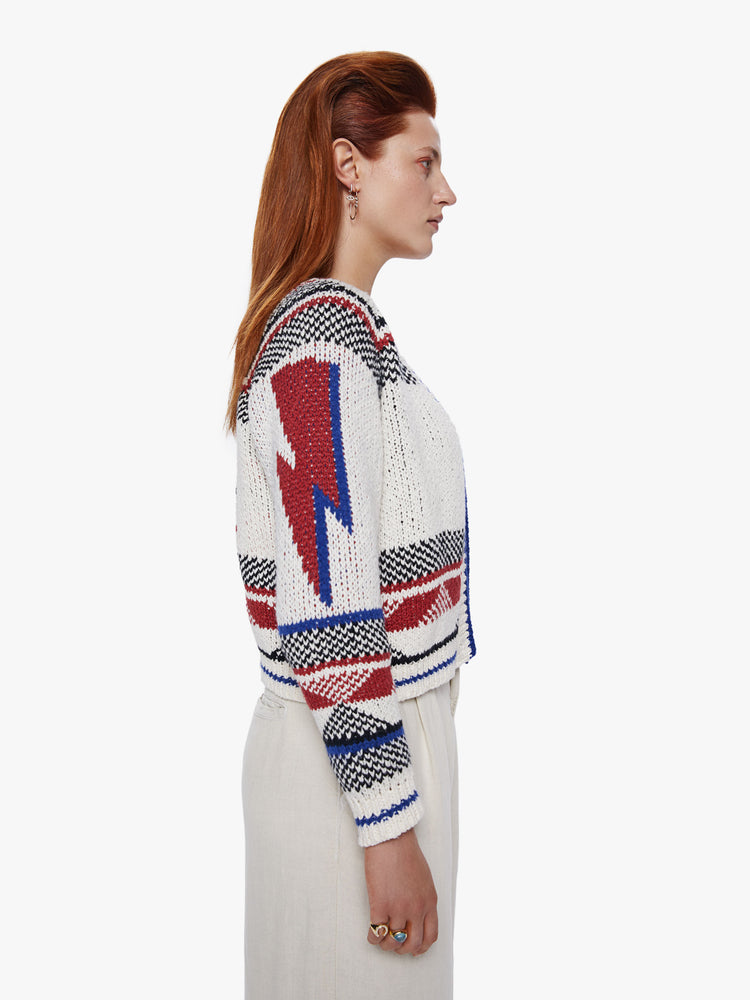 Side view of a woman in a chunky knit cardigan, designed with raglan sleeves, cropped ribbed hems and deep Vneck with three buttons at the waist made from a blend of cotton and alpaca featuring lightning bolts, bold text and knit details in red, blue and black
