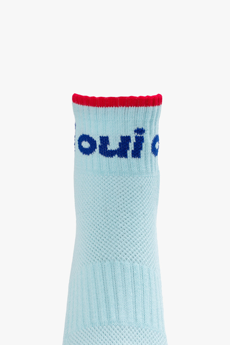 Front close up view of a pair of light blue socks featuring a red stripe and "OUI" in navy blue.