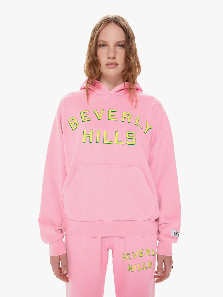 Womens front body view of a bright pink pullover hoodie sweatshirt with a front kangaroo pocket and slightly oversized fit featuring a yellow puff print reading "BEVERLY HILLS"