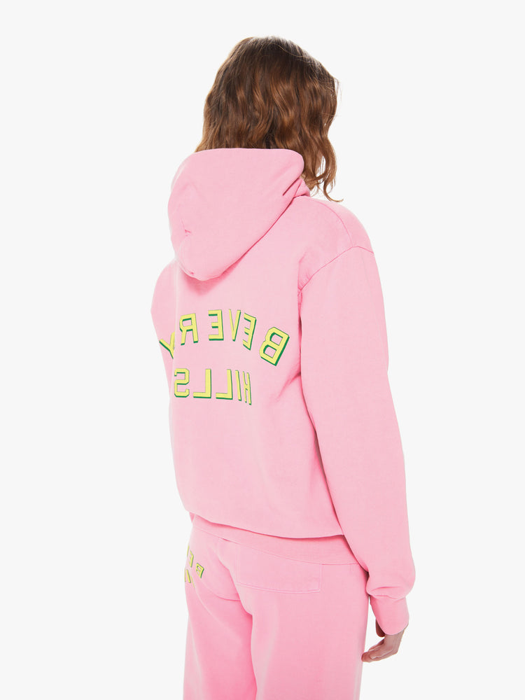 Womens body back 3/4 view of a bright pink pullover hoodie sweatshirt with a front kangaroo pocket and slightly oversized fit featuring a yellow puff print reading "BEVERLY HILLS" in reverse.