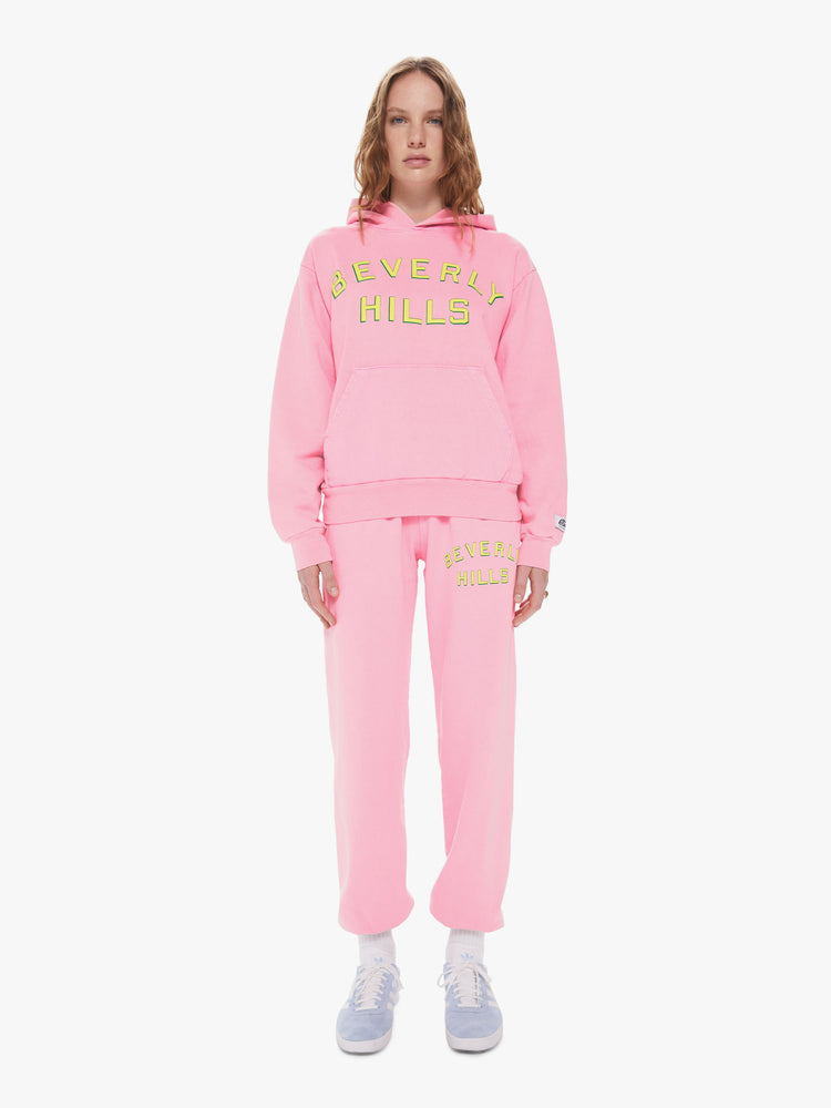 Womens front full body view of a bright pink pullover hoodie sweatshirt with a front kangaroo pocket and slightly oversized fit featuring a yellow puff print reading "BEVERLY HILLS" paired with matching sweatpants.