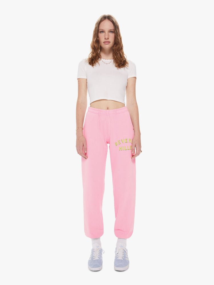 Womens front full body view of a pair of bright pink sweatpants with a slightly oversized fit and elastic waist and cuffs, featuring a yellow puff print on the left thigh reading "BEVERLY HILLS"