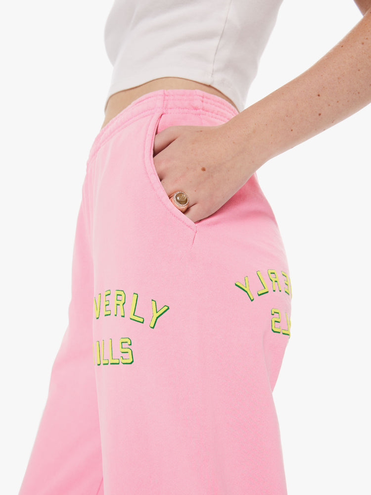 Womens close up view of a pair of bright pink sweatpants with pockets, featuring a yellow puff print on the front and back left thigh reading "BEVERLY HILLS"