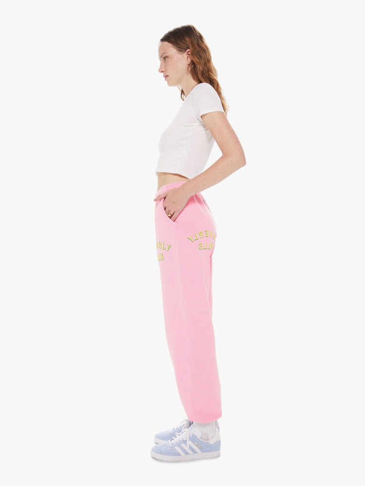 Womens side profile full body view of a pair of bright pink sweatpants with a slightly oversized fit and elastic waist and cuffs, featuring a yellow puff print on the front and back left thigh reading "BEVERLY HILLS" 