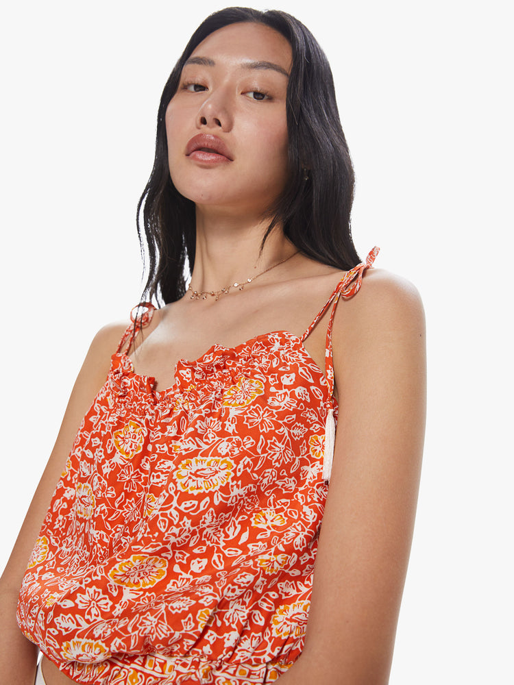 Front close up view of a woman wearing a bright orange top featuring a yellow and white floral print, thin tie straps, and a cropped elastic body.