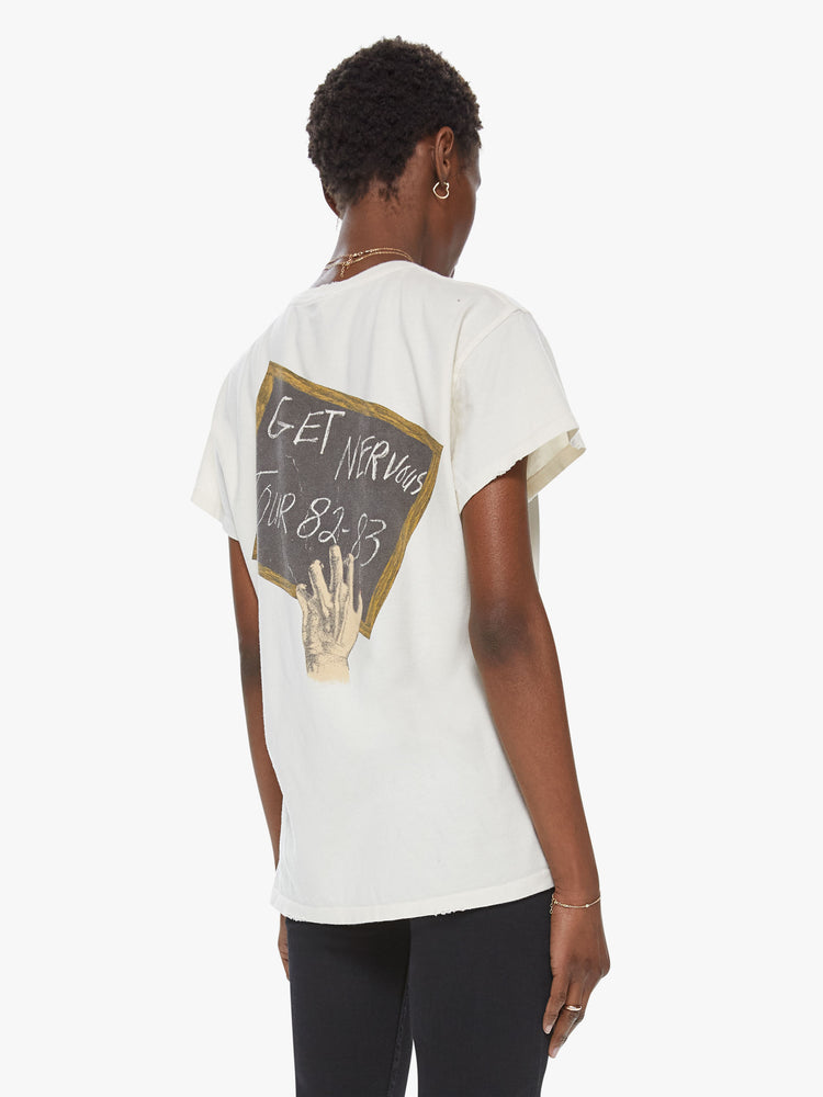 Back view of a woman wearing an off white crew neck tee featuring a vintage band inspired concert graphic on the back reading "GET NERVOUS TOUR 82-83" with distressed details.-