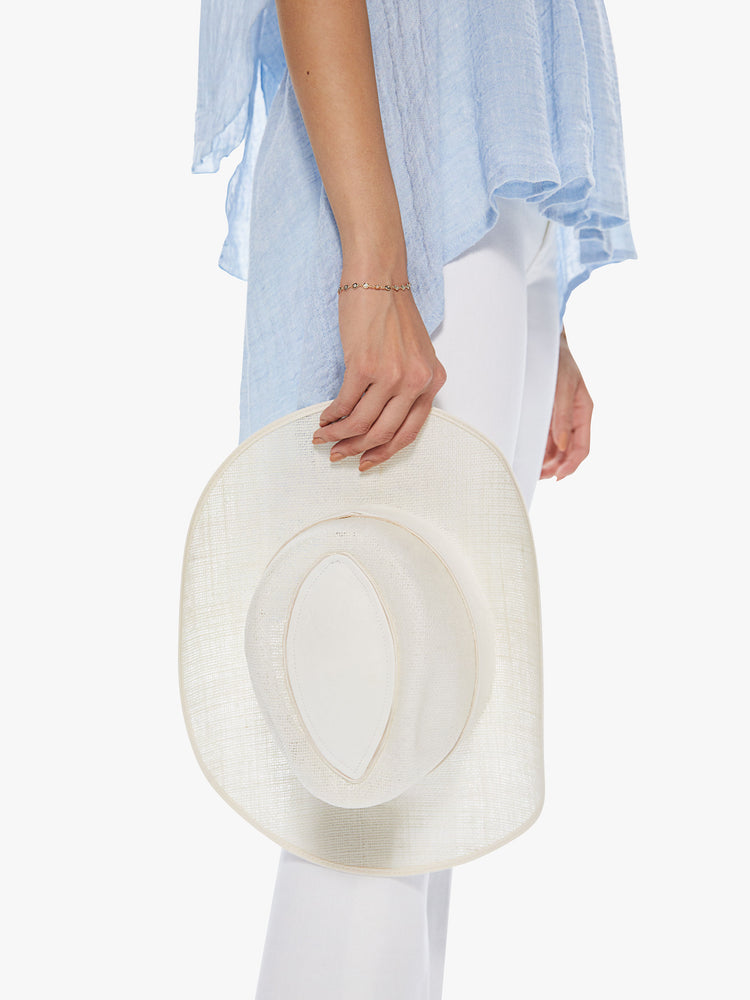 Side view of a woman holding a white linen hat featuring a western inspired shape and leather trim.