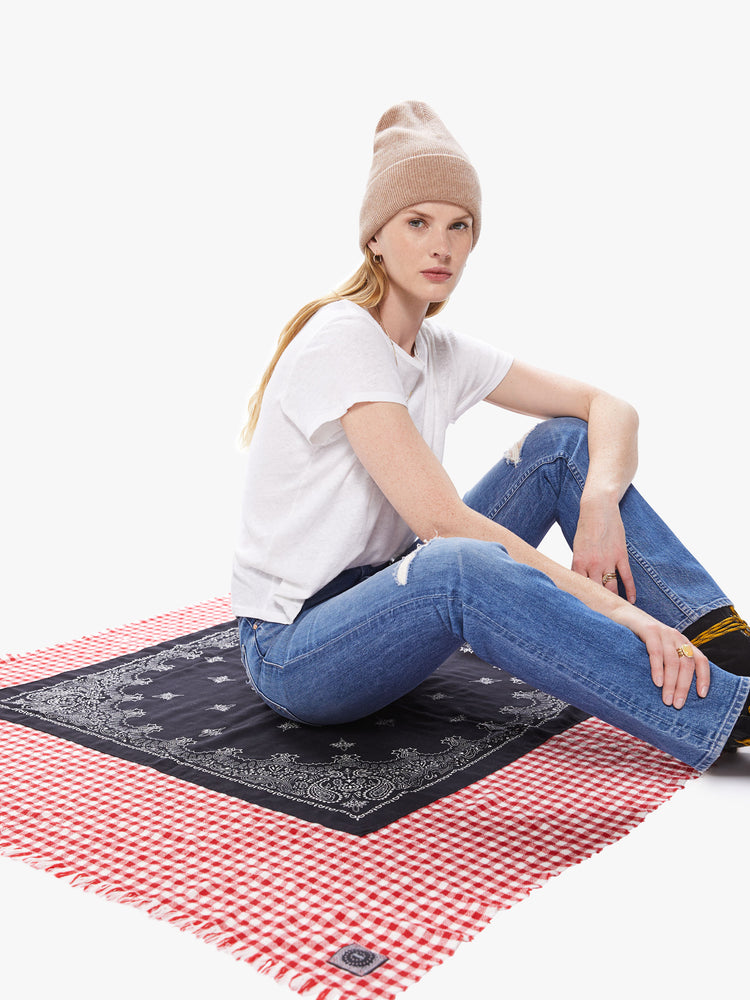 View of a woman sitting on a large square scarf featuring a black bandana print printed over a red gingham print and a fringe hem.
