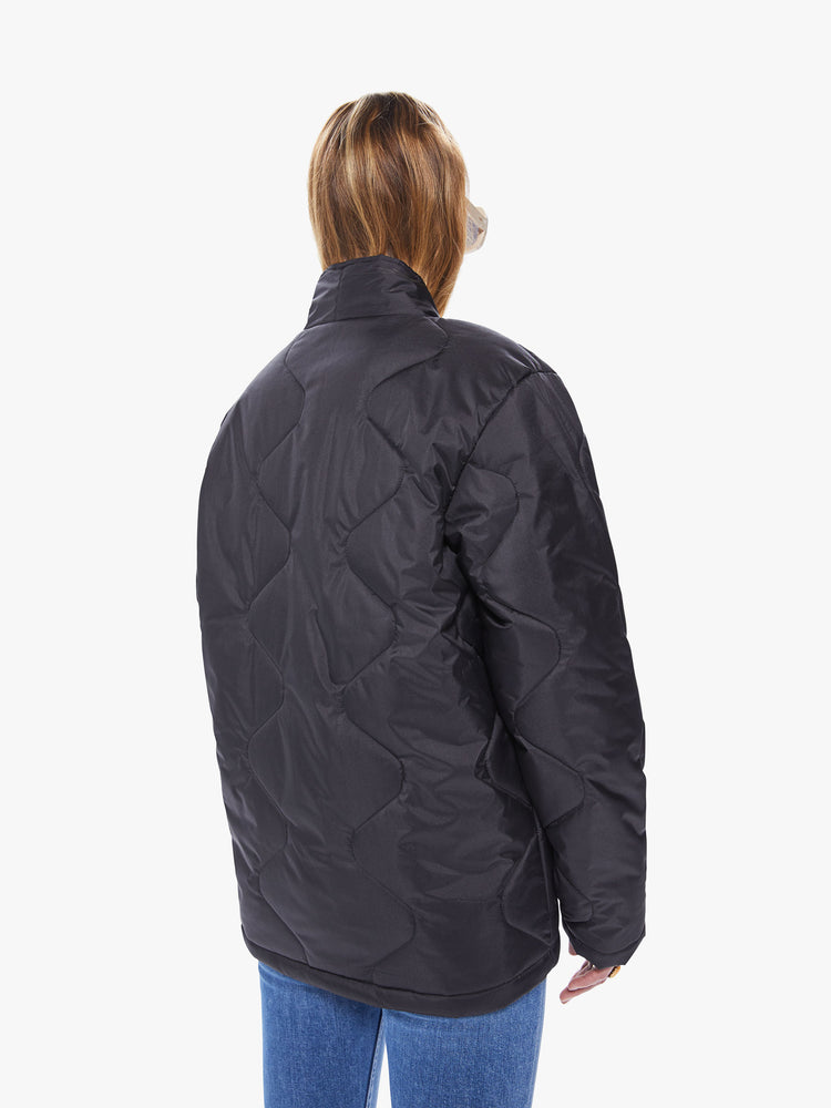 Back view of a women's black quilted jacket, featuring a curved v-neck and front pockets with silver snap buttons and an oversized fit.
