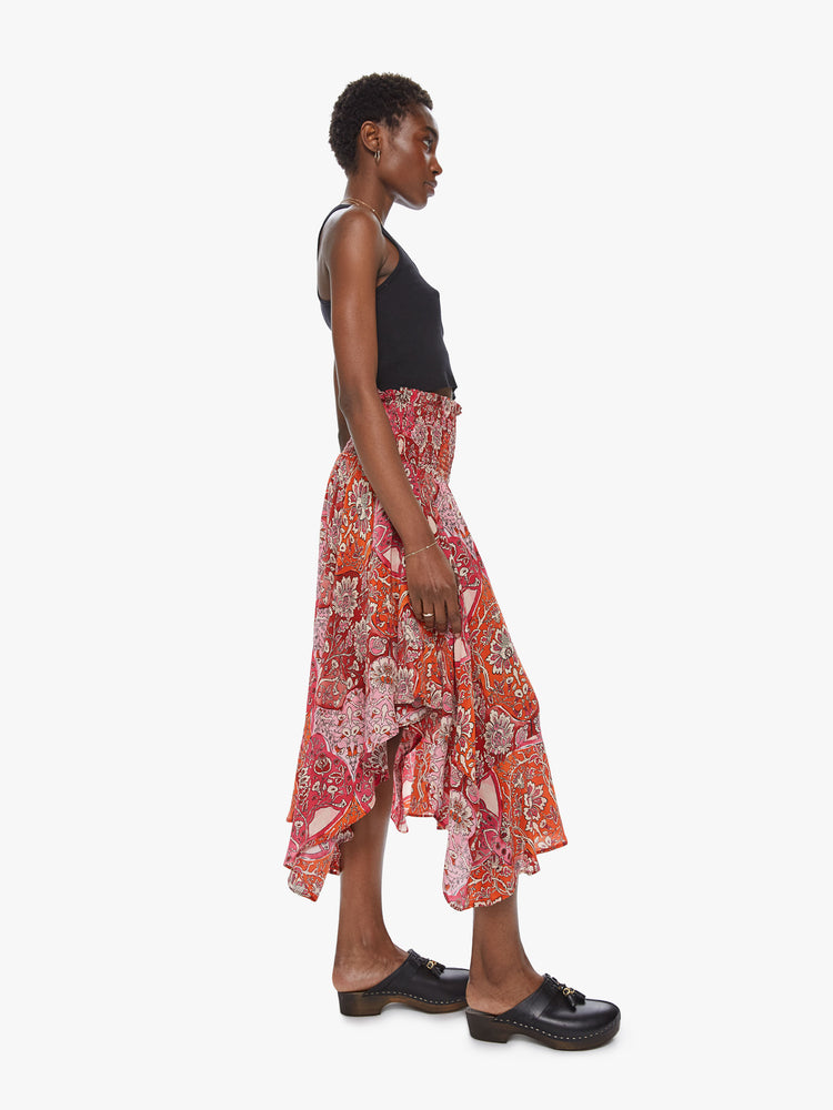Side view of a woman wearing a flowy skirt featuring a red and orange print, a high elastic waist, and an uneven ruffled hem.