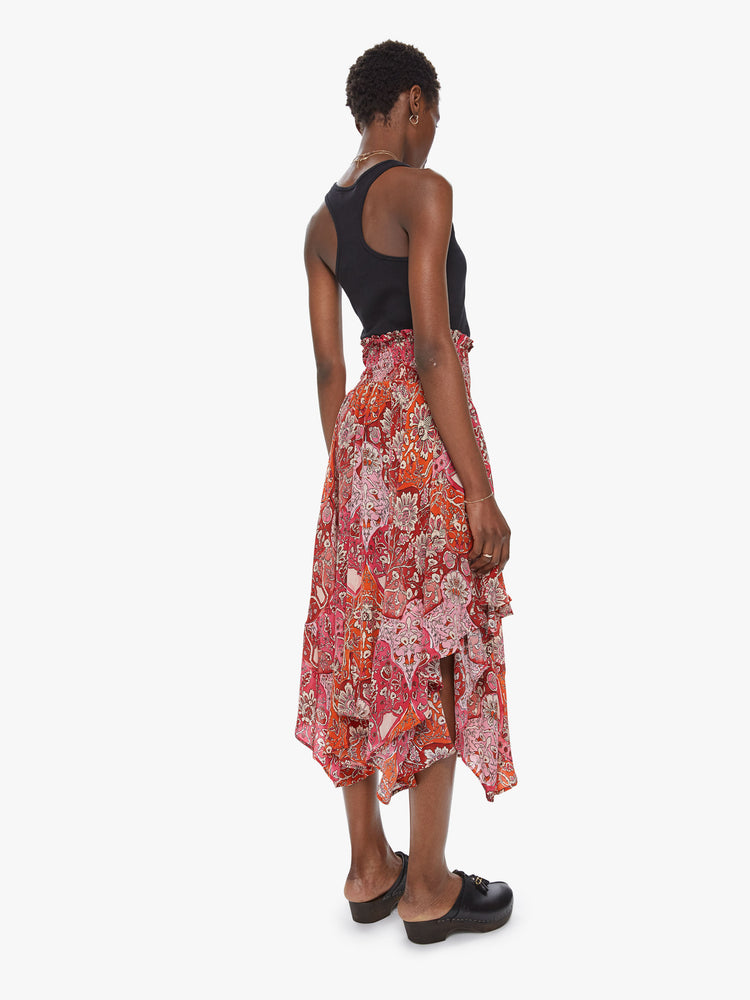 Back view of a woman wearing a flowy skirt featuring a red and orange print, a high elastic waist, and an uneven ruffled hem.