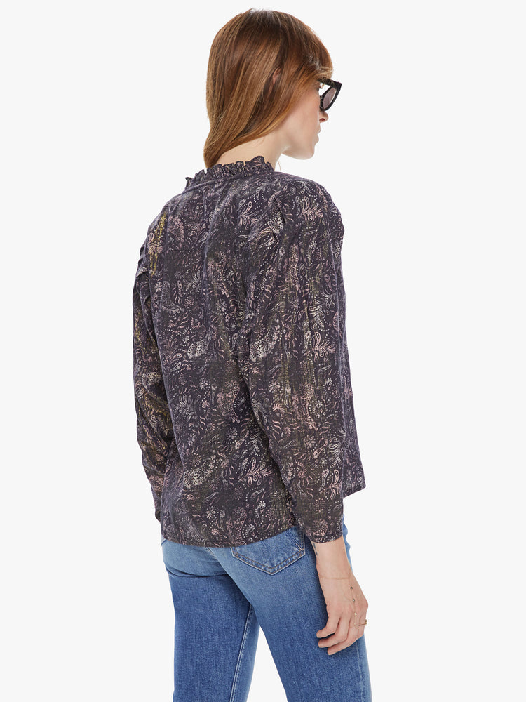 Back view of a woman wearing a long sleeve blouse featuring a dark paisley print with a gold shimmer, ruffled sleeves, and a tie vneck.