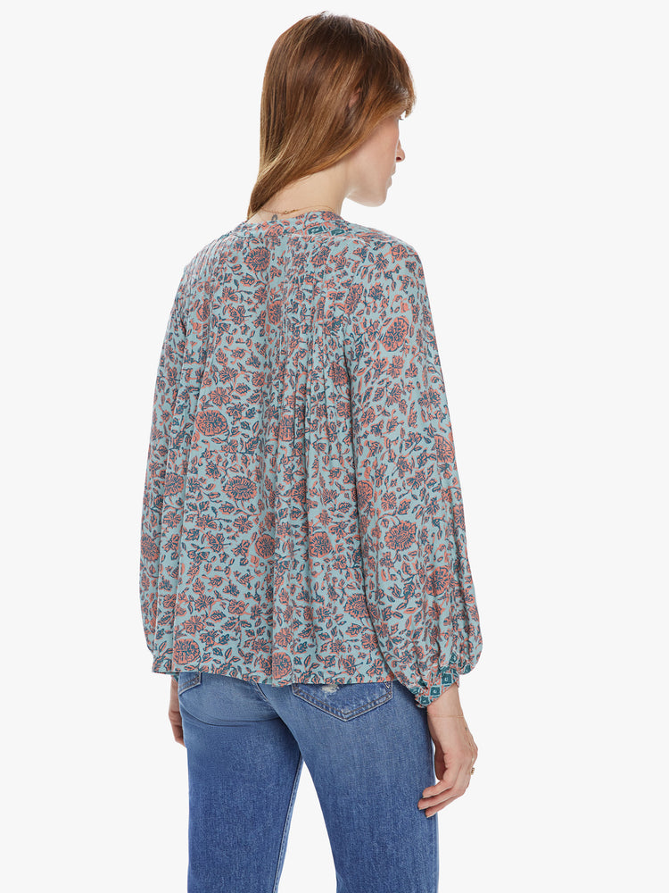 Back view of a woman wearing a flowy top featuring long sleeves, a deep v-neck with a tie, and a pale green and coral floral print.