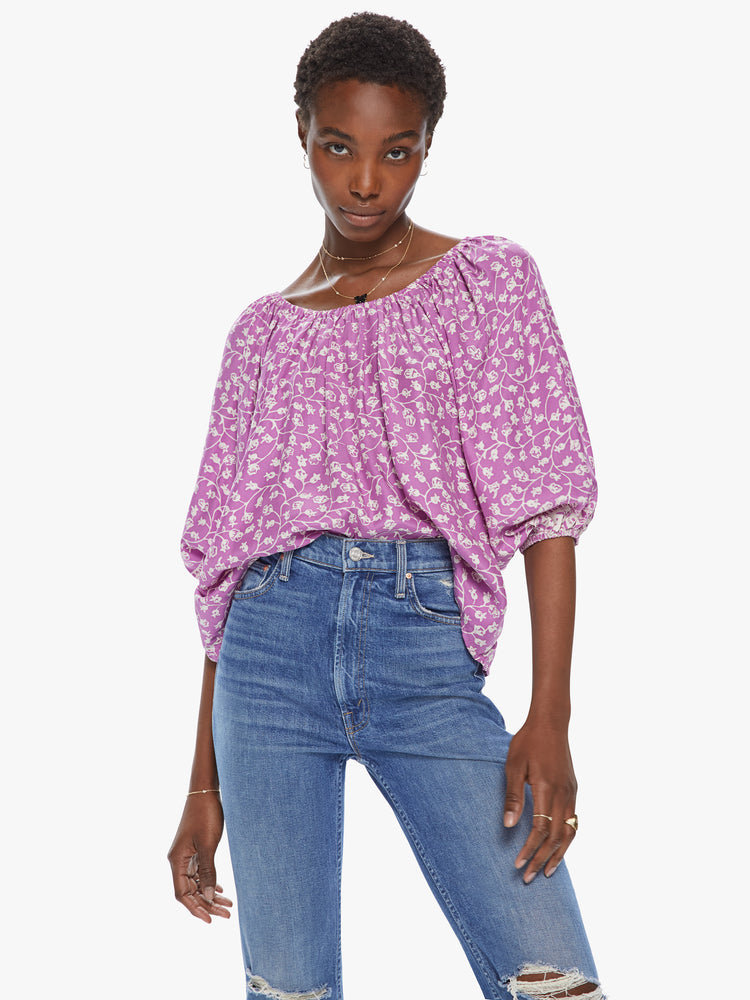 Front view of a woman wearing a flowy top featuring a wide elastic neck, half length billow sleeves, and a purple and white floral print.
