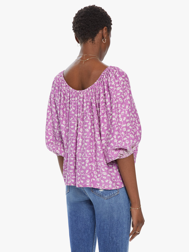 Back view of a woman wearing a flowy top featuring a wide elastic neck, half length billow sleeves, and a purple and white floral print.