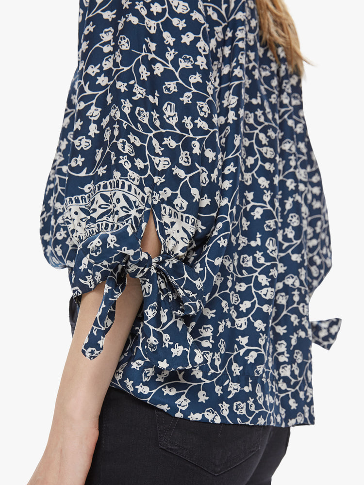 Side close up view of a woman wearing a flowy blouse featuring 3/4 length sleeves with a tie, a buttoned v neck, and an indigo floral print.