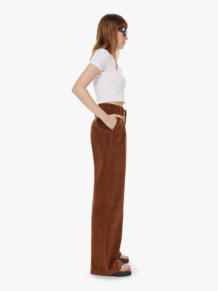 Side view of a woman wearing dark brown corduroy pants featuring a high, pleated waist and wide straight legs.