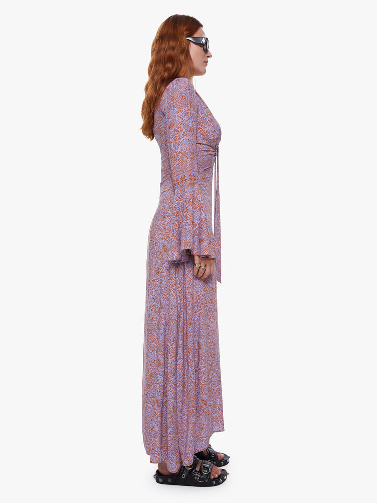 A side view of a woman wearing an orchid-purple and rust floral print, full length dress with bell sleeves and keyhole neckline.